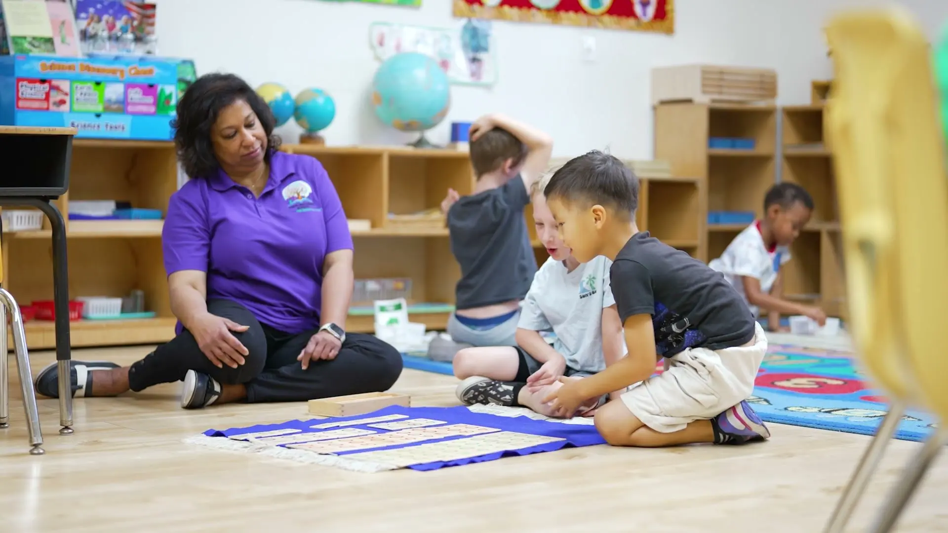 A teacher in a purple school shirt sits on the classroom floor while boys work with Montessori number materials.