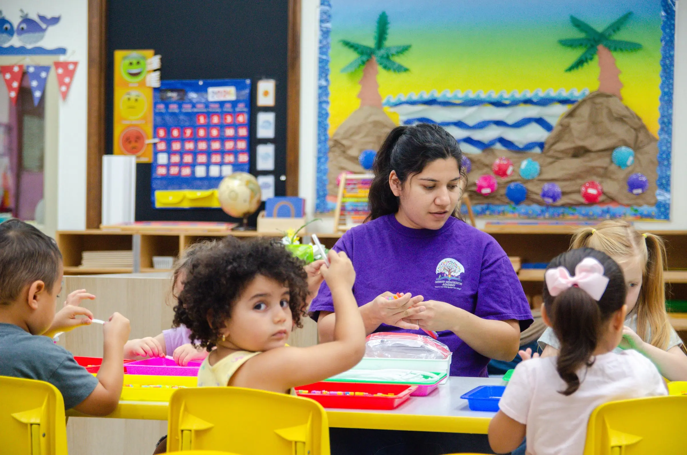 A teacher in a purple school shirt works with children at a table covered in colorful trays.