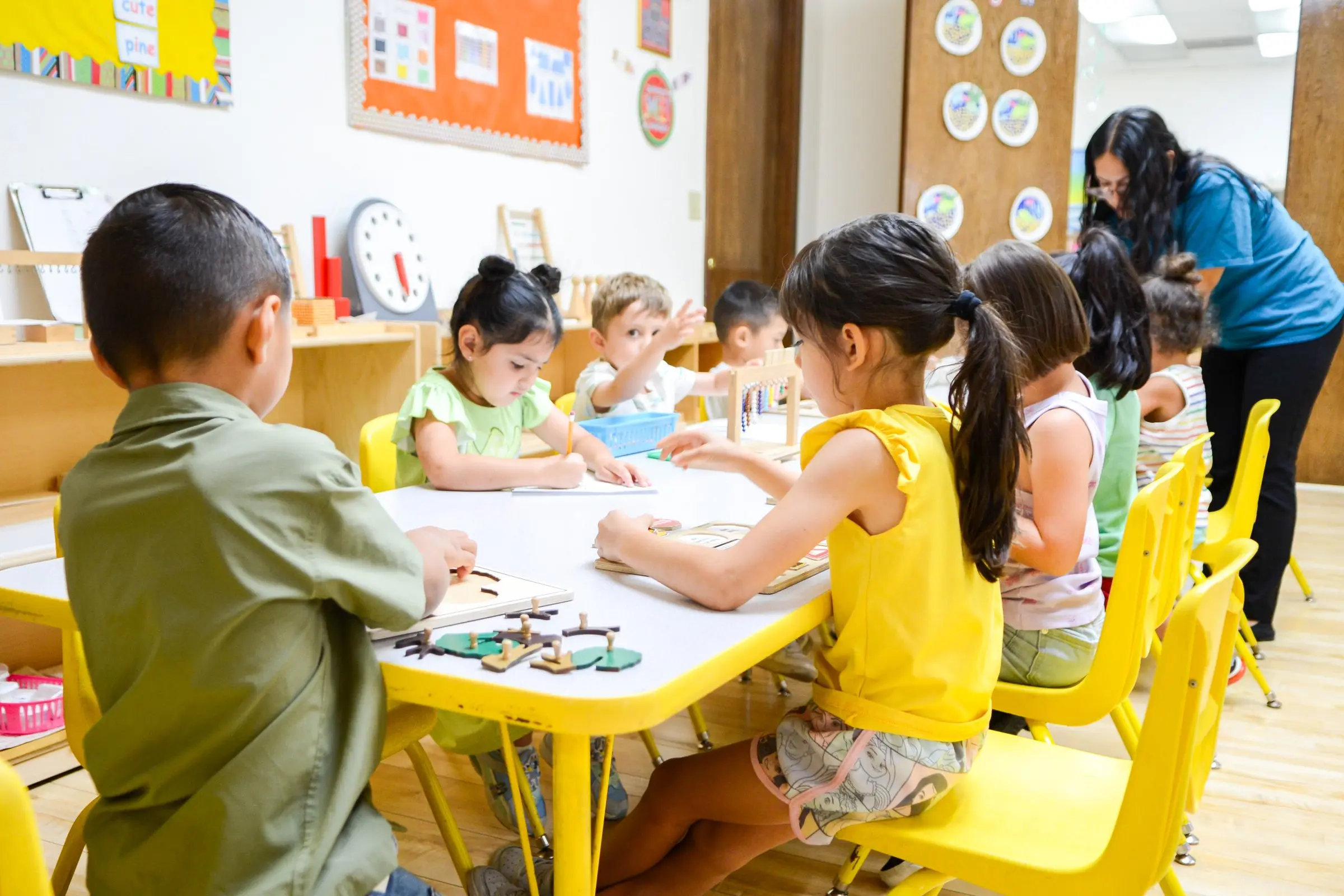 Children work at their tables with wooden puzzles while a teacher helps nearby.