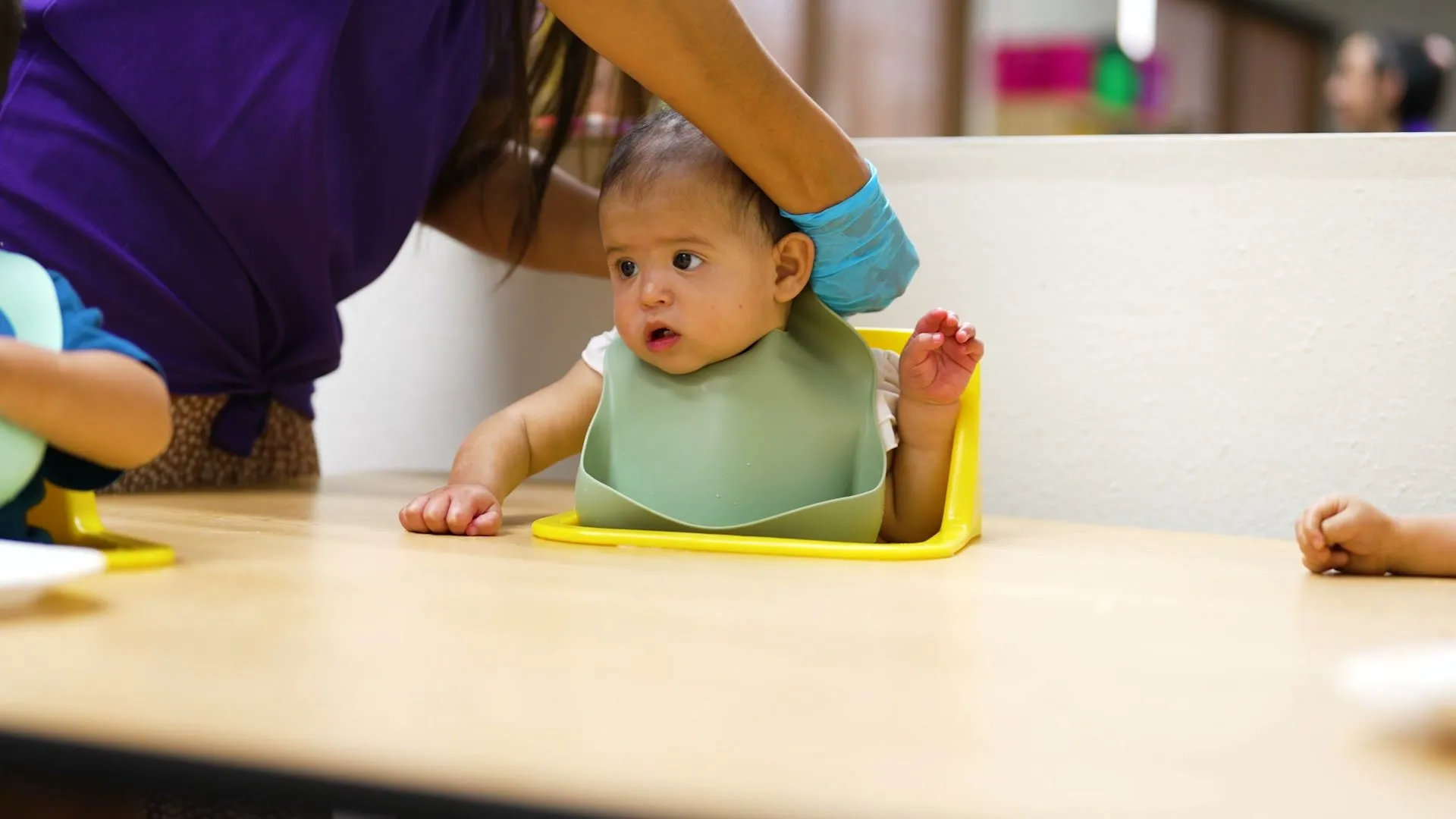 A baby in a bib sits in a feeding chair at the table while a teacher helps her settle in.