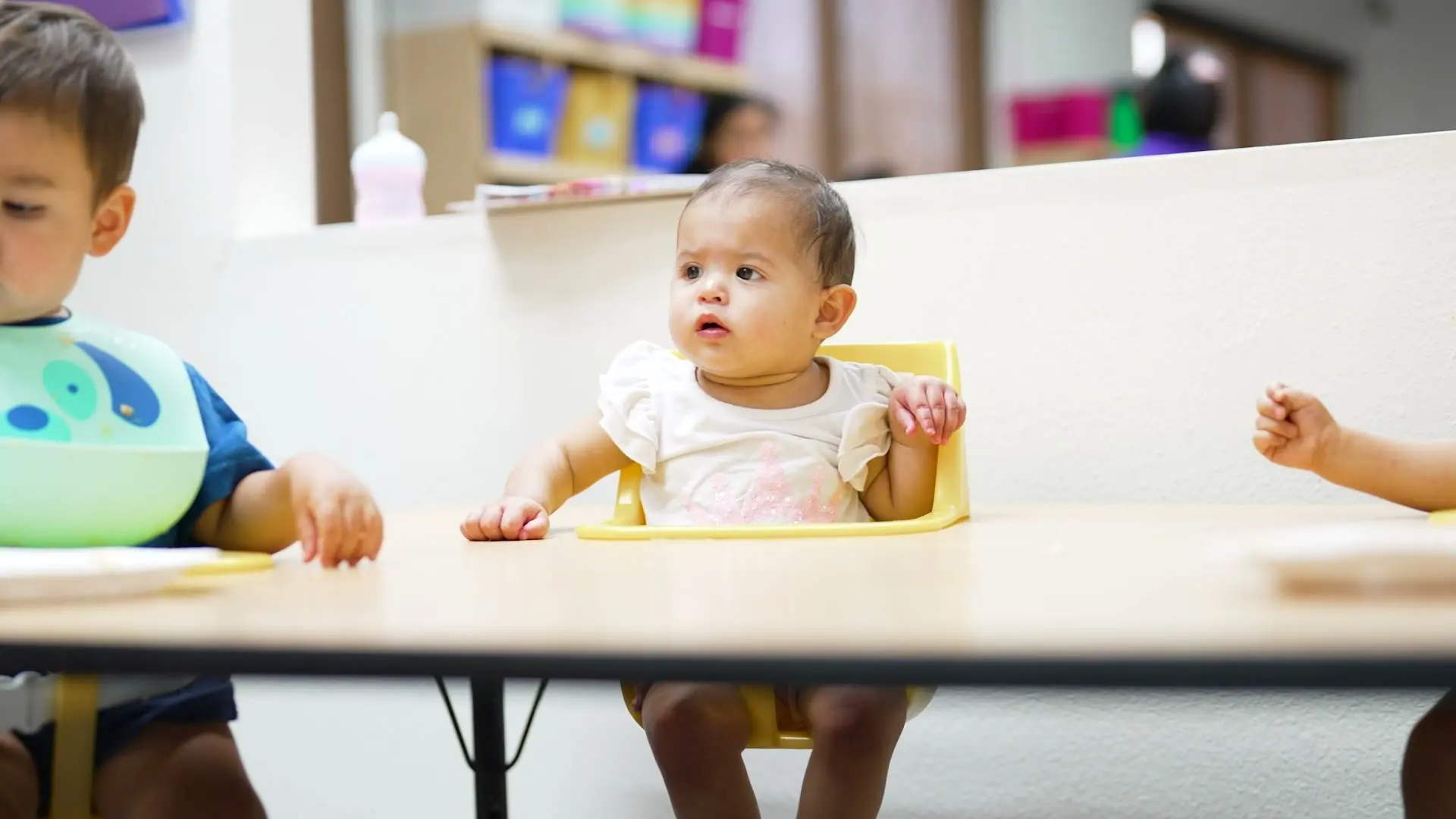 A baby sits in a small chair at the table in the infant room, looking up.
