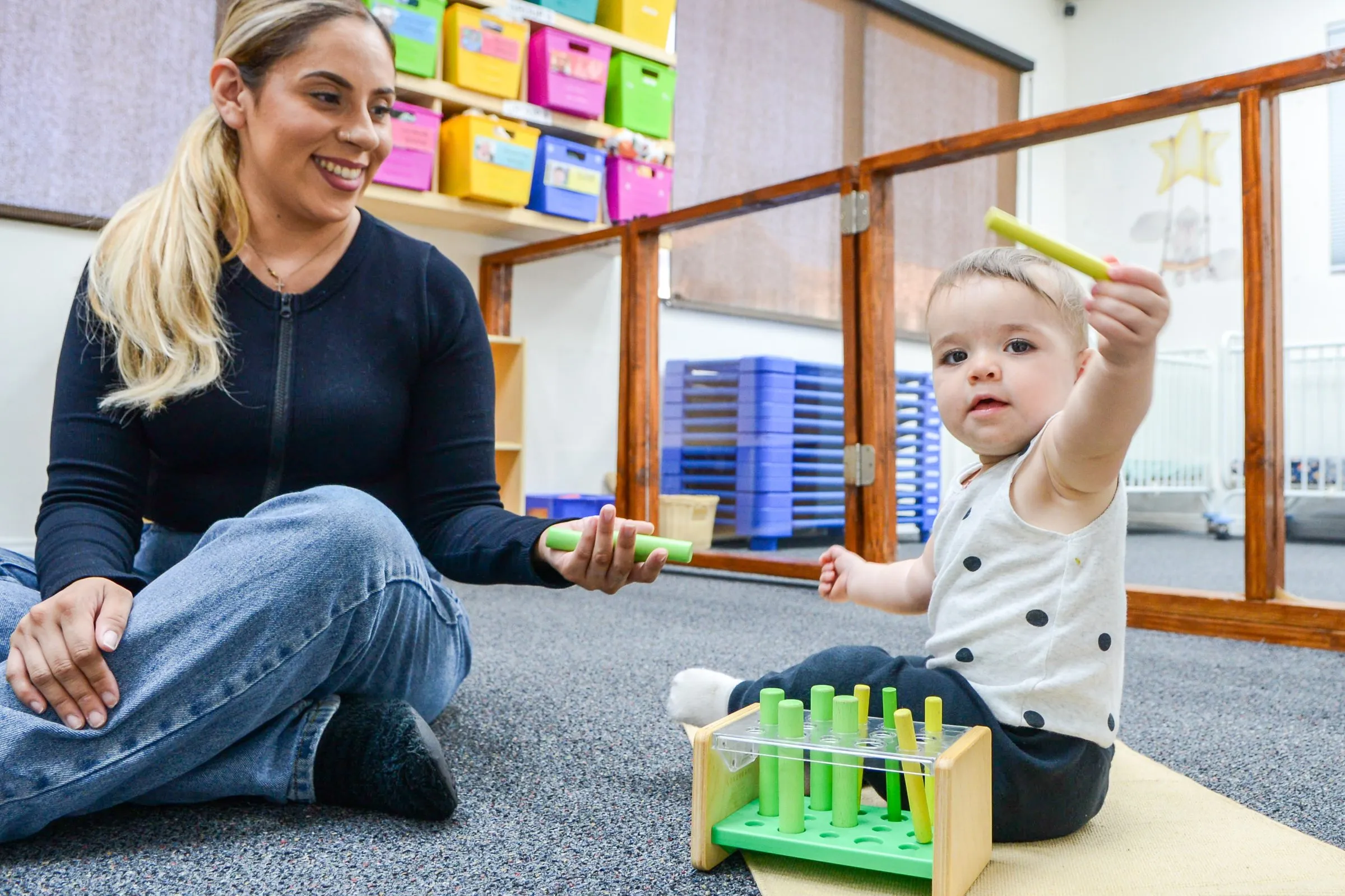 A smiling teacher sits on the carpet as a baby holds up a toy peg.