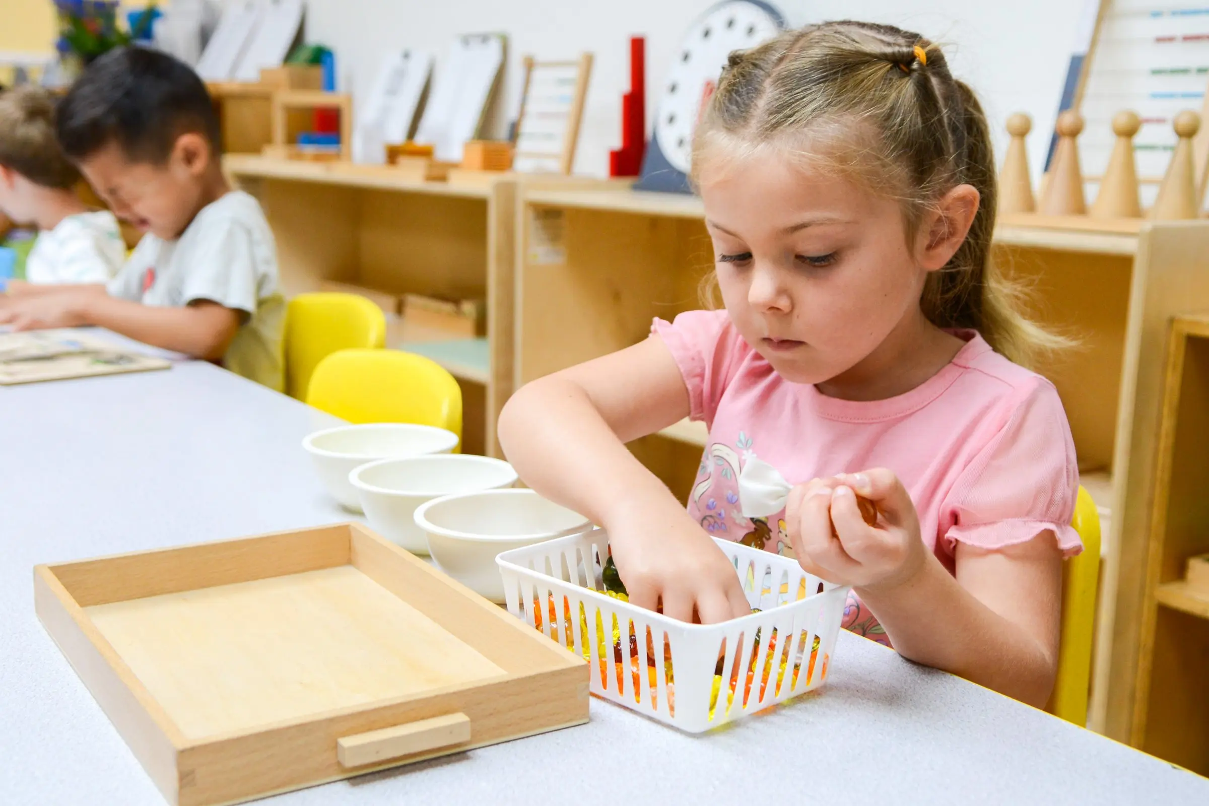 A girl uses tongs to sort small objects into a basket at a Montessori shelf.