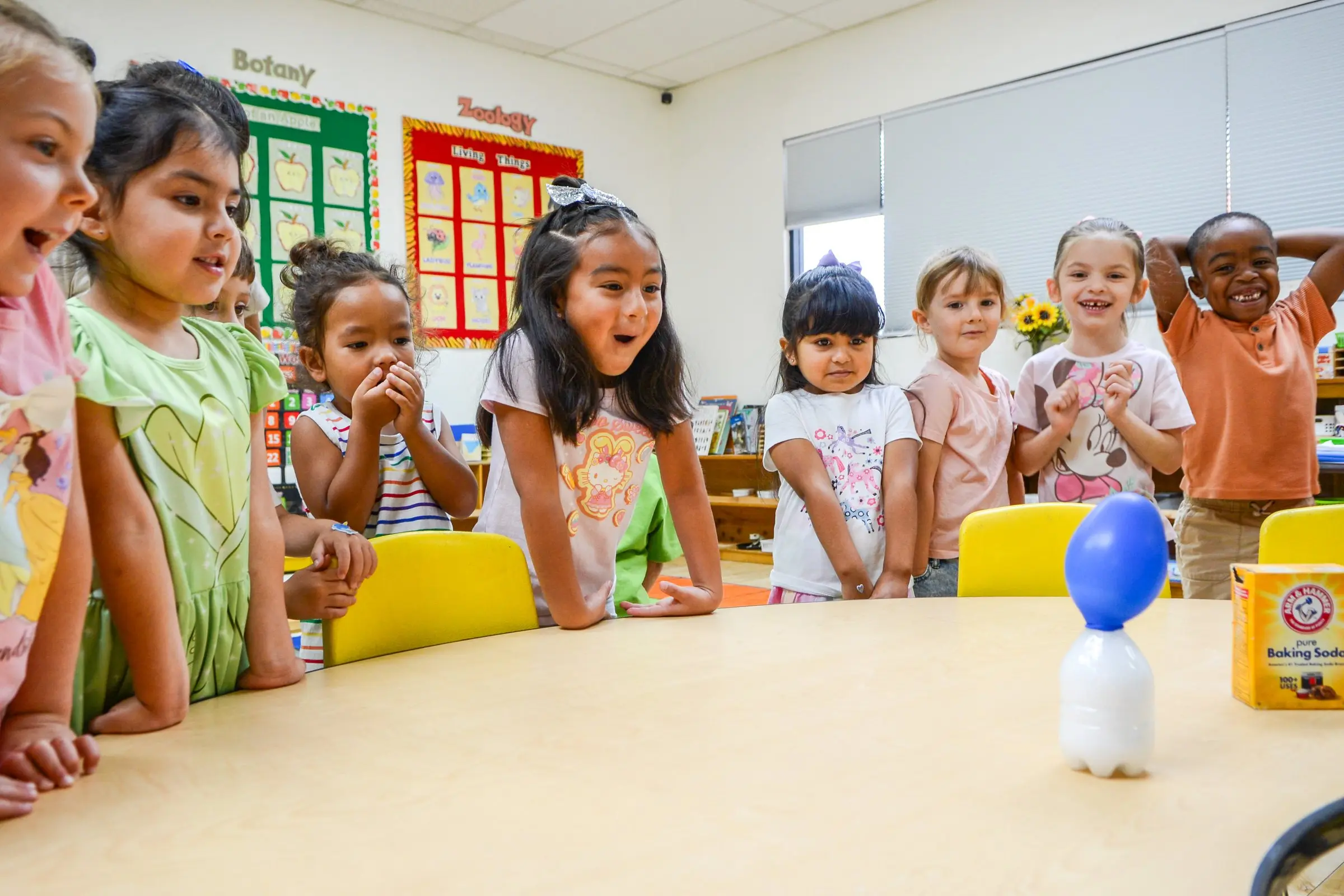 Children crowd around a table with wide eyes, watching a science experiment with a balloon.