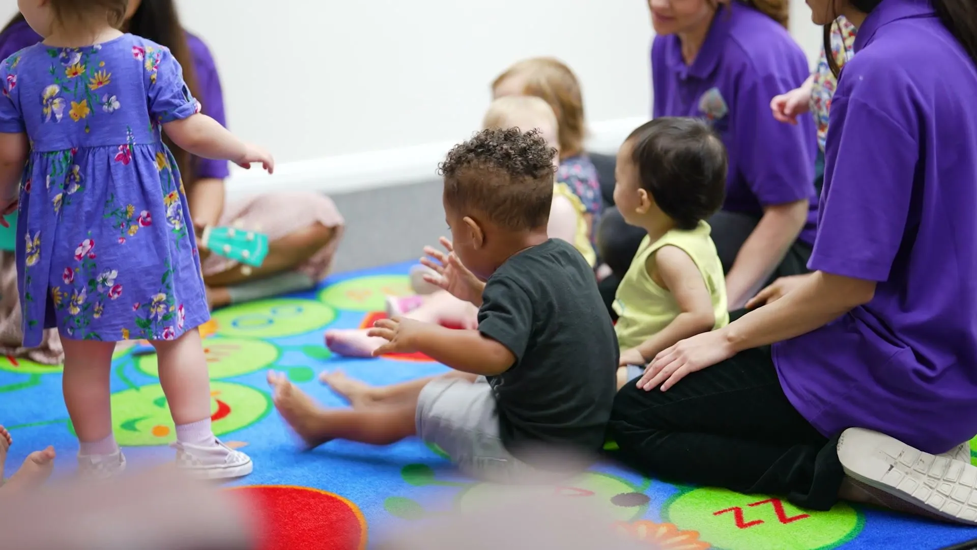 Toddlers sit on a colorful rug with their teachers during a group activity.
