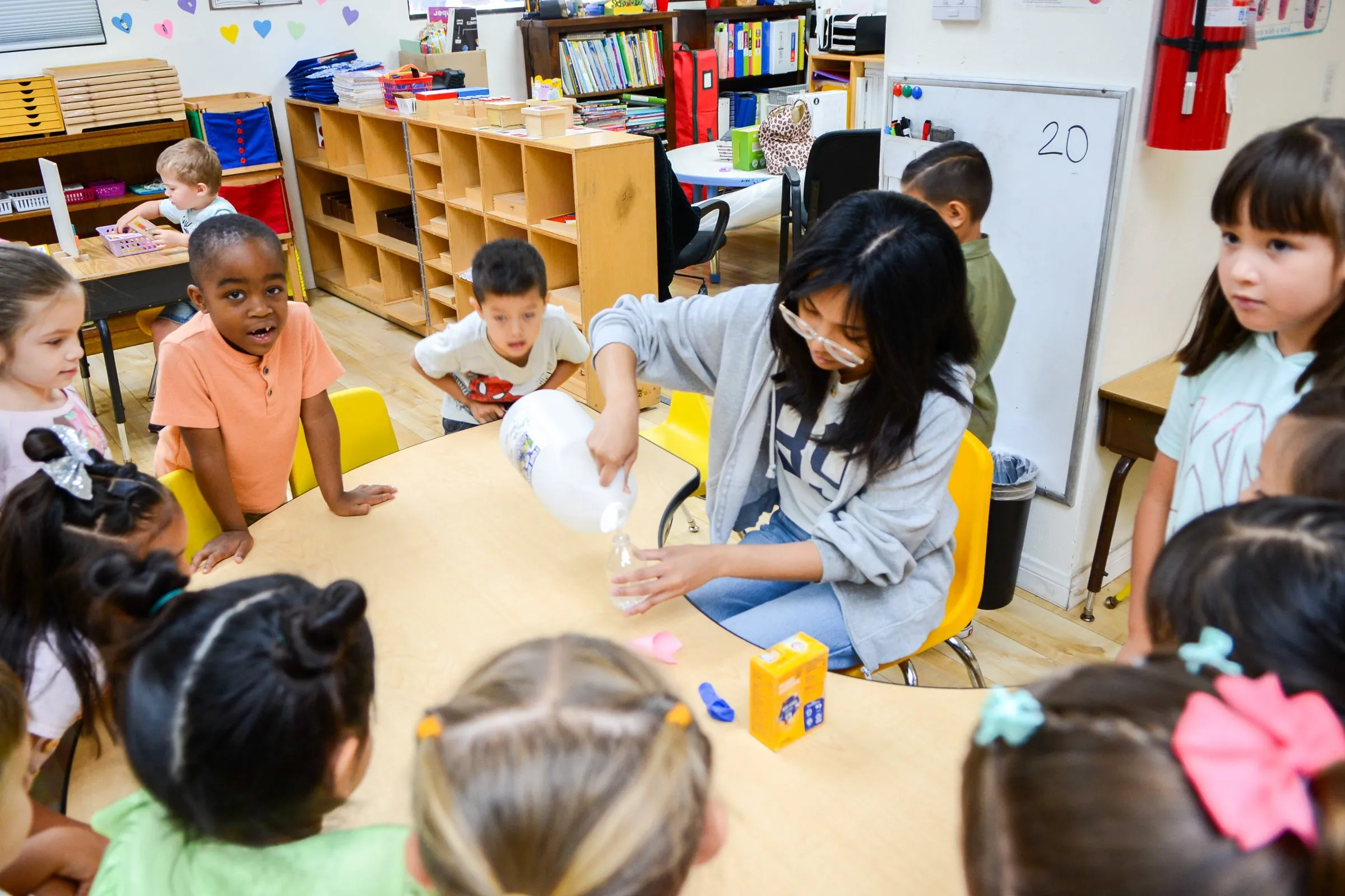 Children gather around a round table as their teacher leads a hands-on science activity.