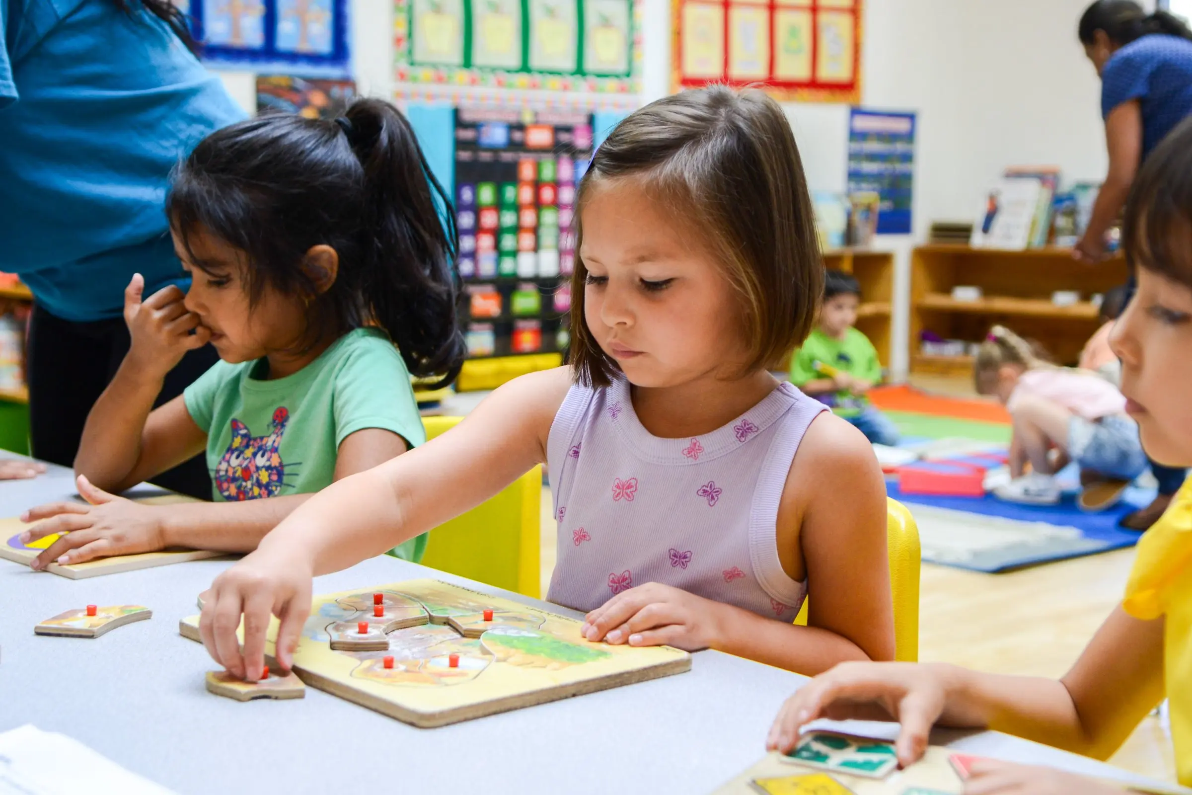 Two girls work on wooden puzzles at a classroom table.