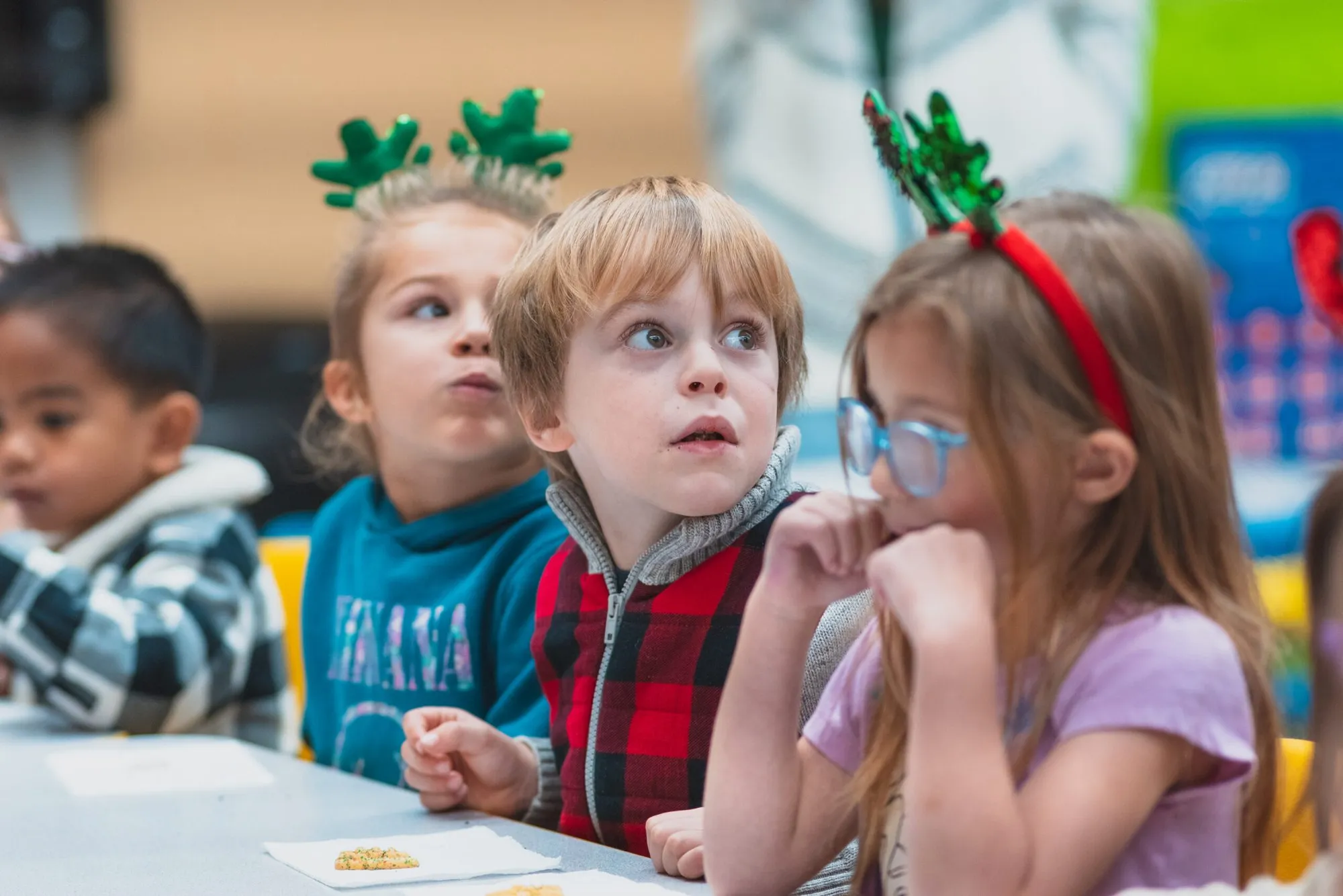 Four children in festive headbands sit at a classroom table during a holiday celebration.