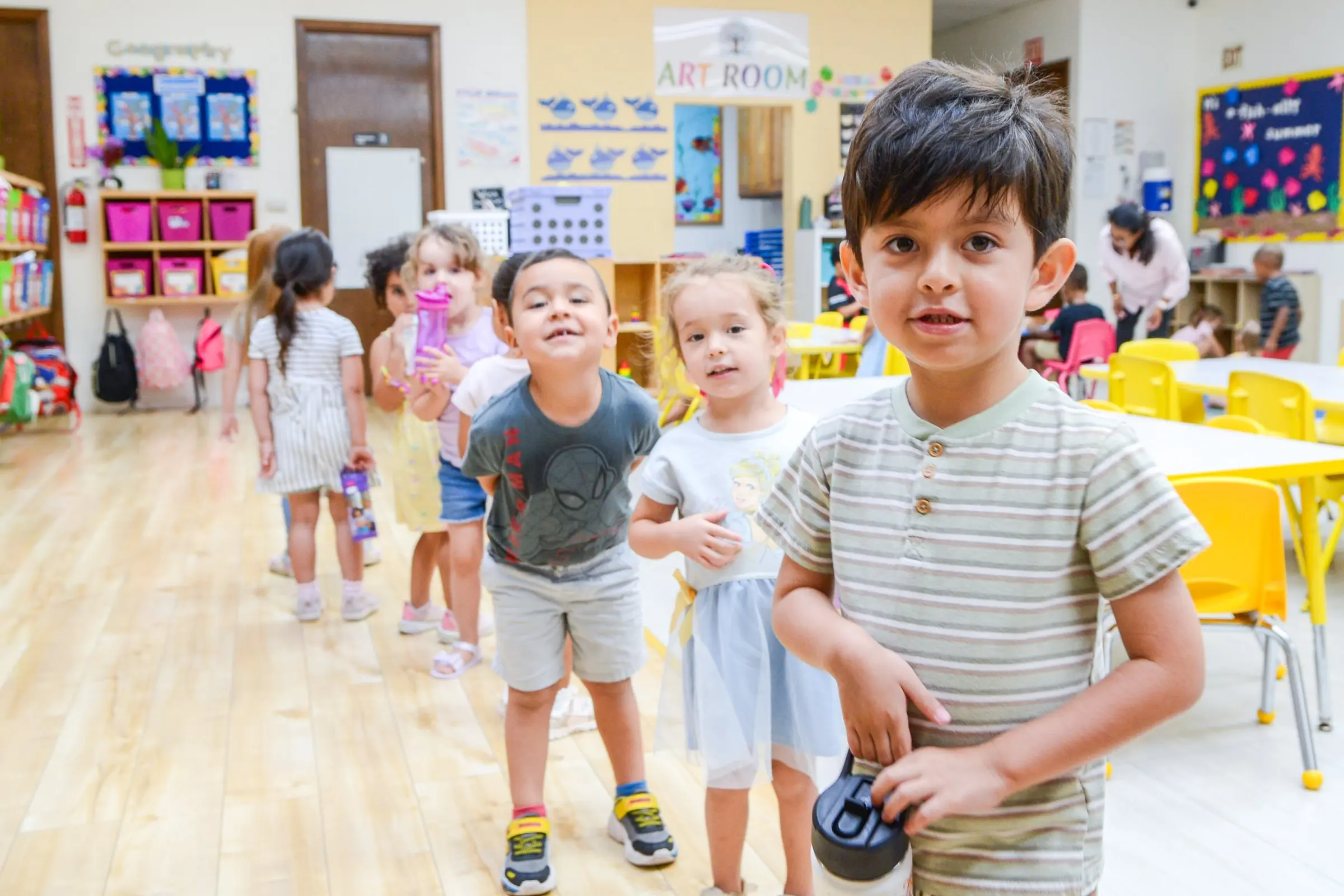 A smiling boy and his classmates walk through a bright classroom.