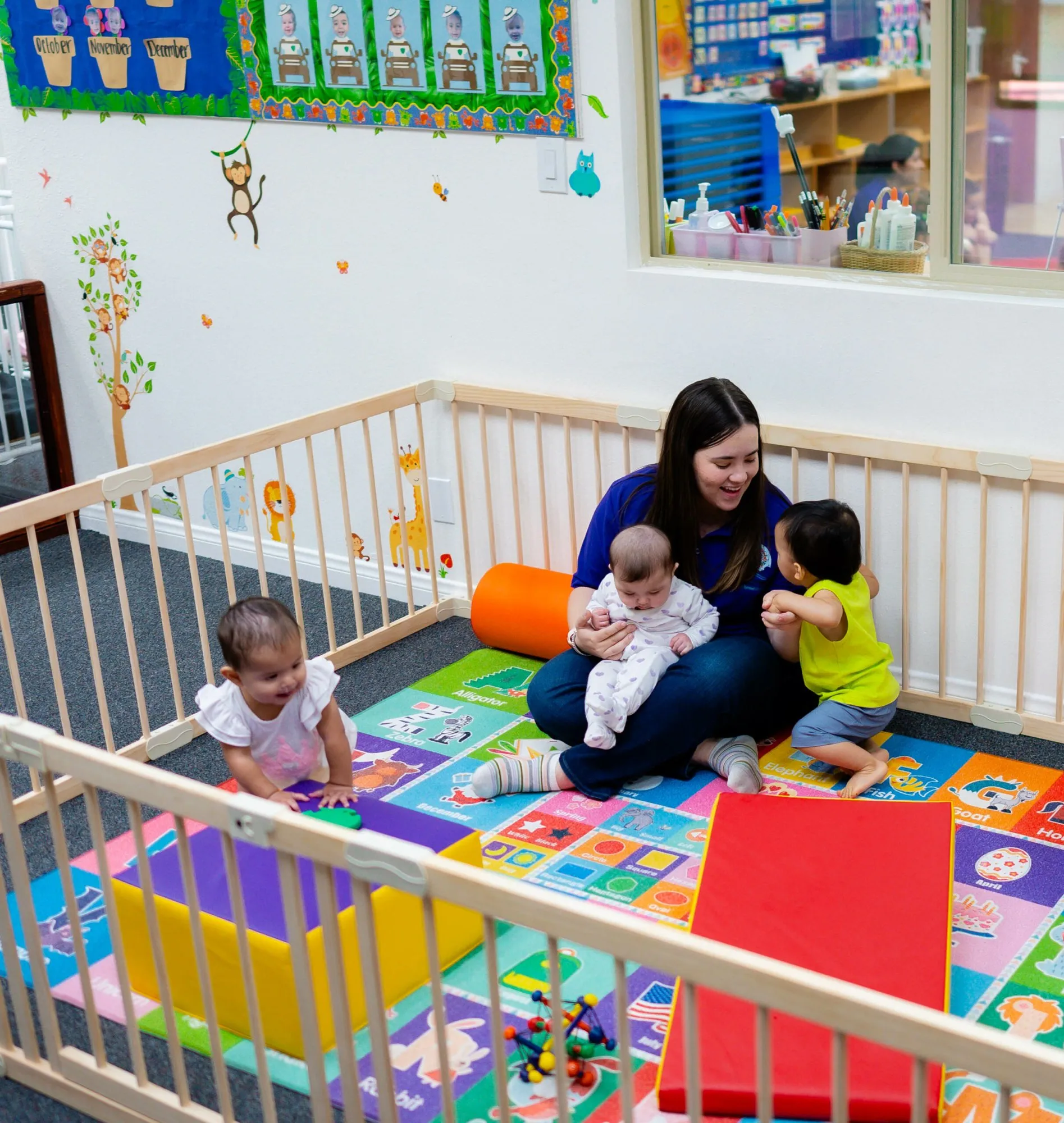 A teacher on a colorful play mat holds a baby while two toddlers play beside her.