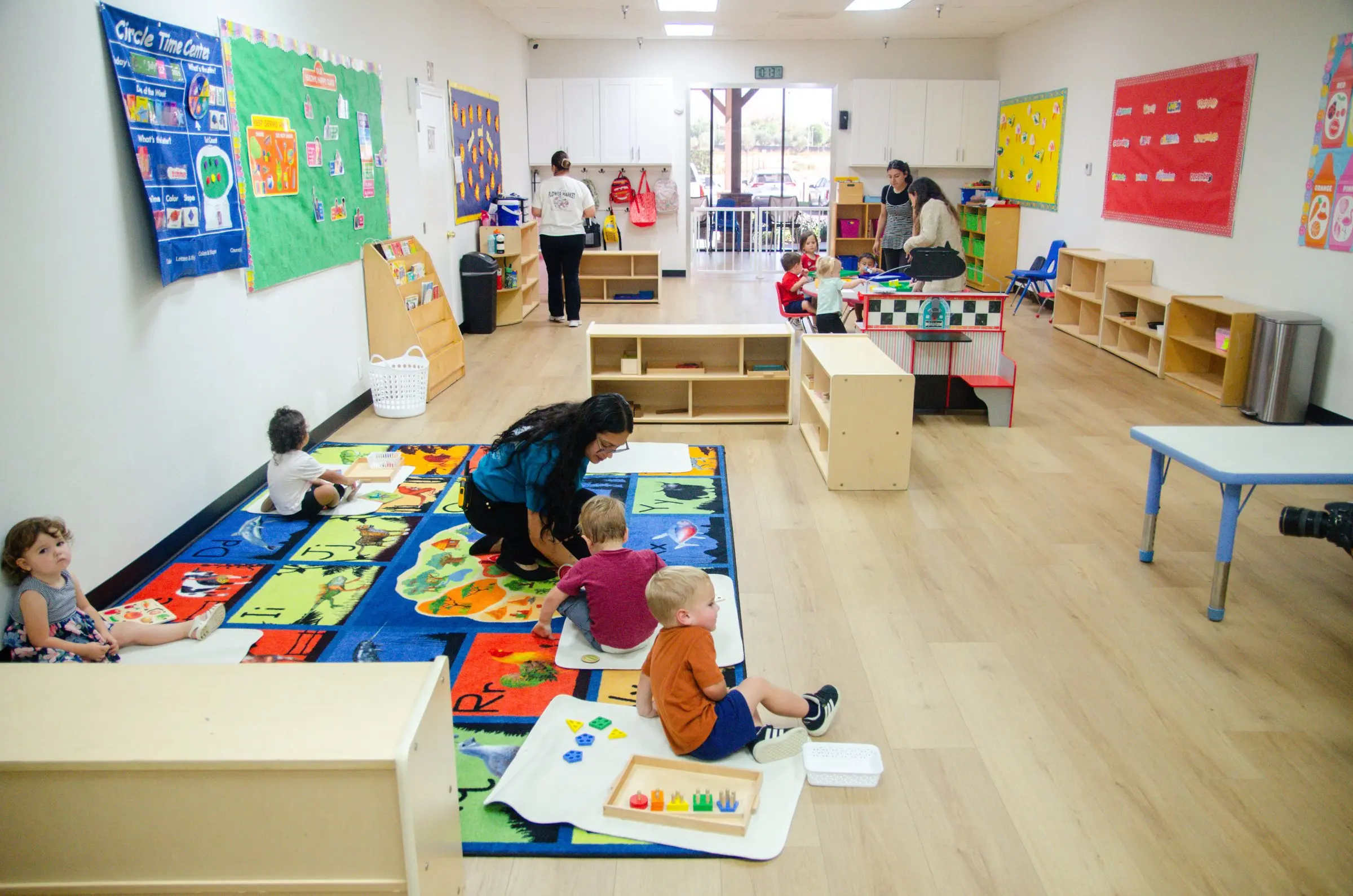 A bright infant and toddler room with a colorful rug, low shelves and a teacher playing with toddlers on the floor.