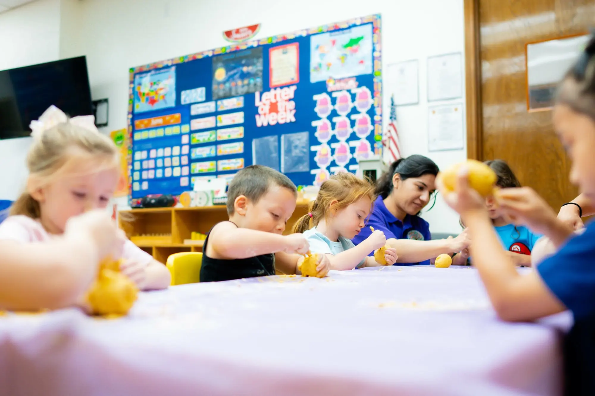 Children squeeze lemons at a classroom table while a teacher helps a classmate beside them.