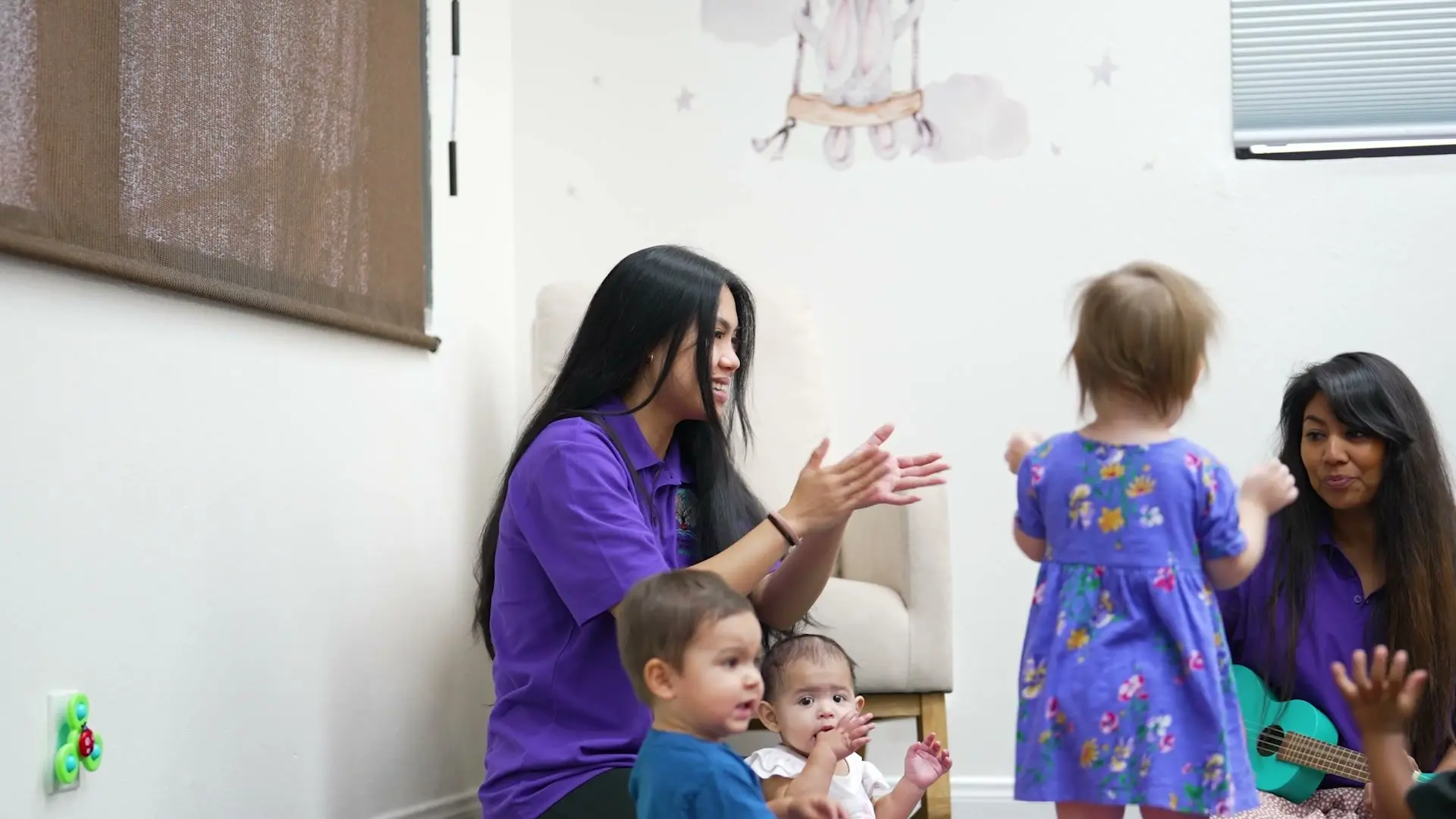 Two teachers sing and clap with a group of toddlers, one playing a ukulele.