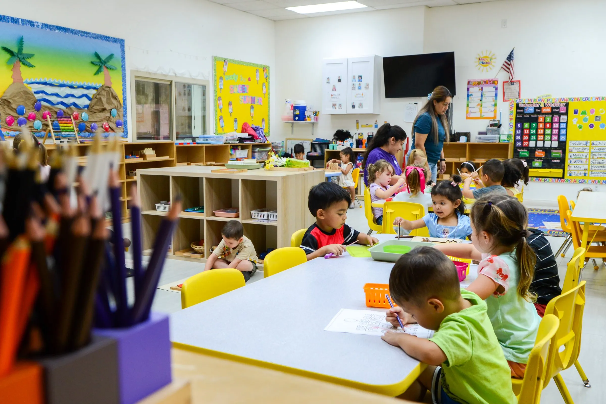Children work at tables in a bright, busy classroom, with colored pencils in the foreground.