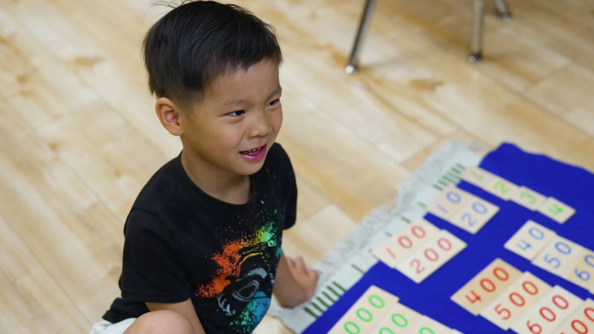 A smiling boy lays out Montessori number cards on a mat on the classroom floor.